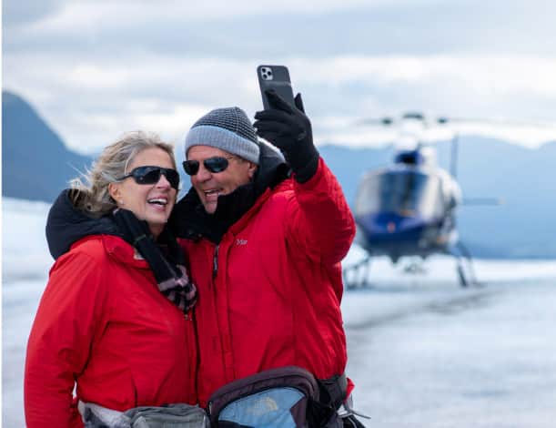 A couple dressed in jacket and gloves take a selfie atop a glacier with a tour helicopter parked behind them